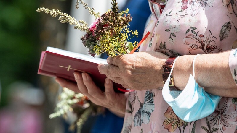 Eine Gottesdienstbesucherin hält in einem Festgottesdienst zu Mariä Himmelfahrt vor der Wallfahrtskirche Maria Eich ihre Kräuterbuschen und ein Gesangsbuch in den Händen. Am Arm trägt sie einen Mundschutz. Traditionell werden zu Mariä Himmelfahrt lebensnotwendige und heilkräftige Pflanzen wie Brotgetreide, Heil- und Gewürzpflanzen zu Kräuterbuschen gebunden und im Festgottesdienst gesegnet.